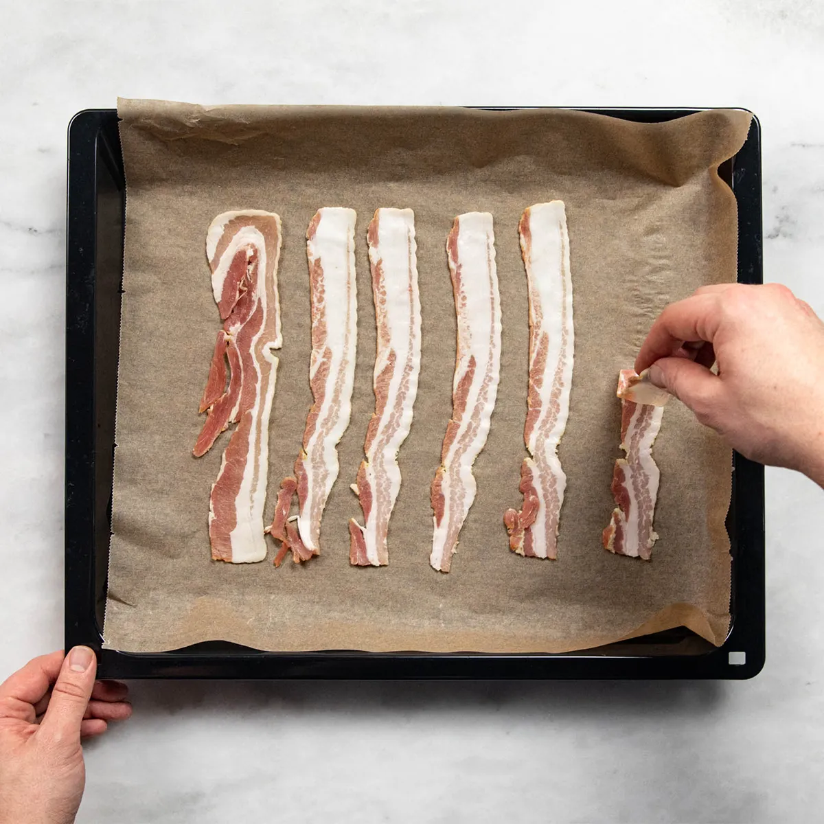 Hands placing raw bacon strips on a parchment-lined baking tray on a marble countertop.