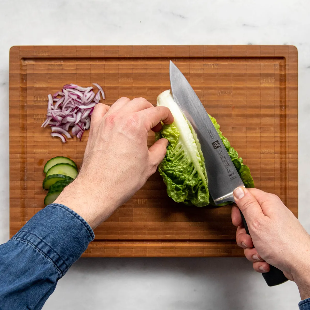Top-down view of two hands slicing a small romaine lettuce with a chef’s knife on a wooden cutting board; sliced red onion and cucumber pieces sit beside it, with a light marble counter and a “kitchen stories” logo in the top right.