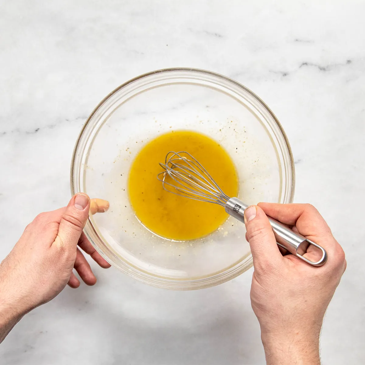Top-down view of two hands whisking a golden vinaigrette in a clear glass mixing bowl on a light marble countertop; a small metal whisk is held in the right hand while the left steadies the bowl, with a “kitchen stories” logo in the top right.