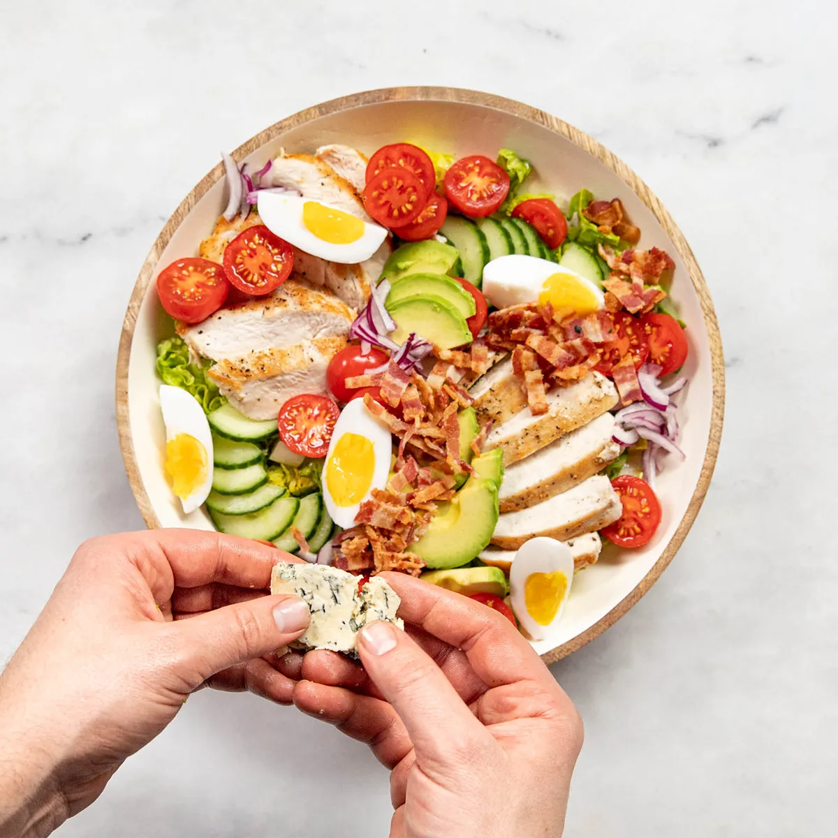 op-down view of a composed salad in a wooden bowl on a light marble surface: sliced grilled chicken, avocado, cucumber slices, cherry tomatoes, quarters of boiled eggs with bright yolks, crumbled bacon and red onion, while hands break a piece of blue cheese over the salad. "kitchen stories" logo in the top-right.