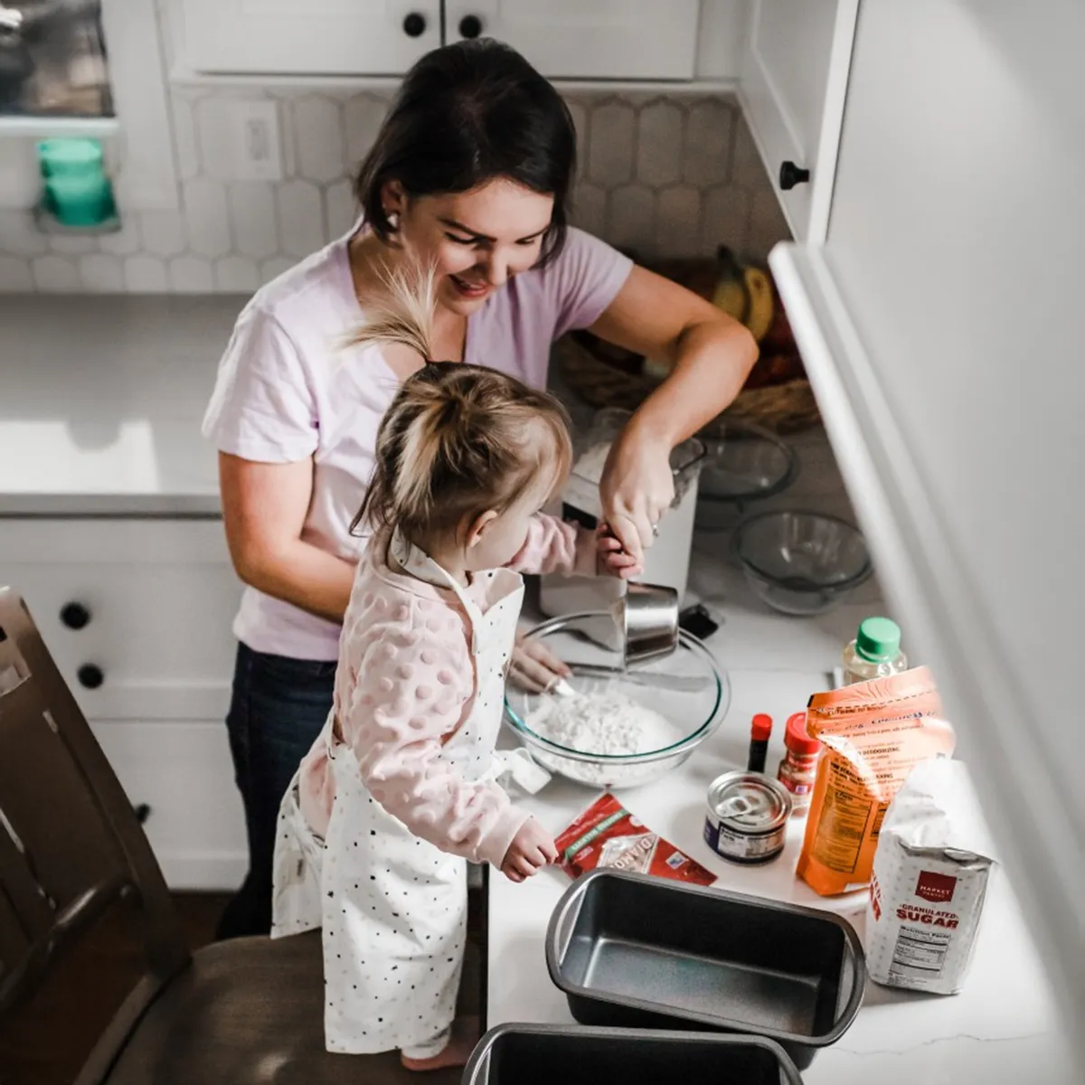 Adult and child in white aprons baking together in a kitchen, pouring ingredients into a glass bowl with baking supplies nearby