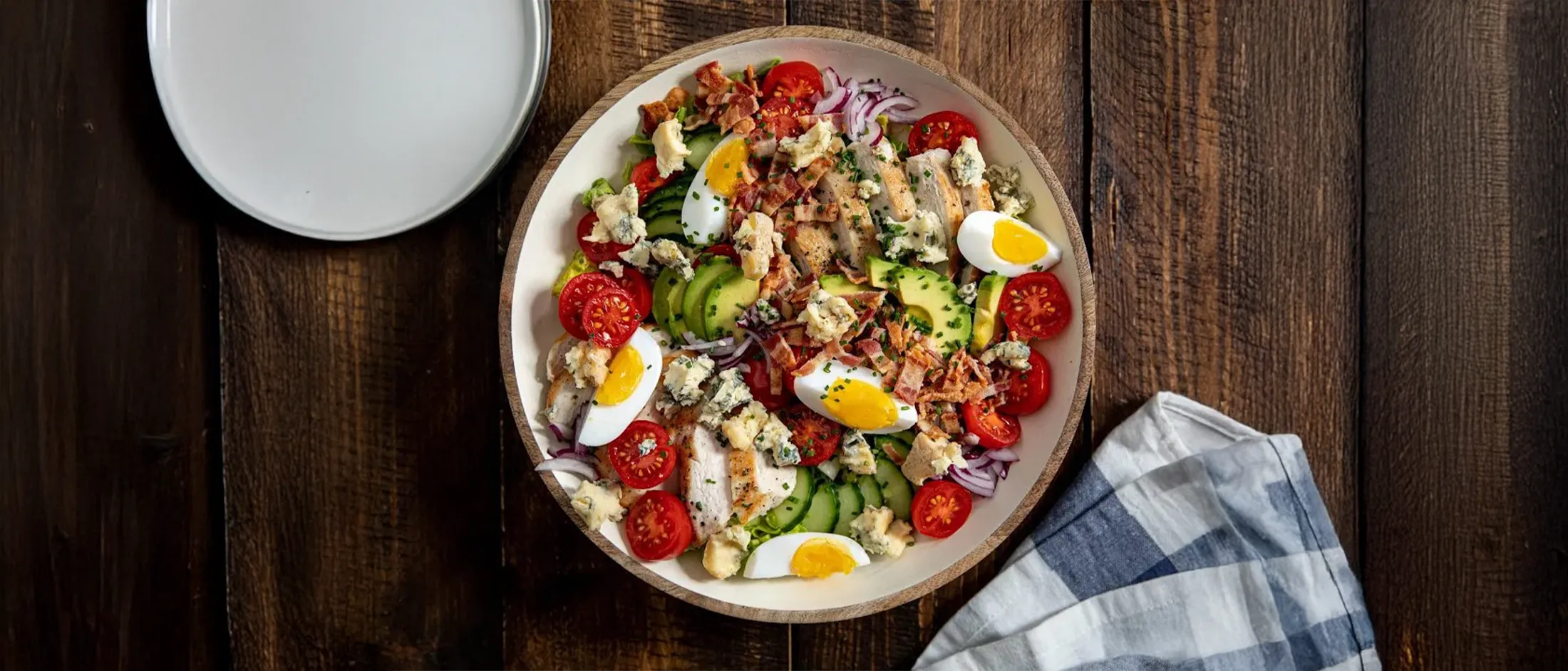 A vibrant Cobb salad with chicken, bacon, avocado, eggs, tomatoes, and blue cheese, on a rustic wooden table.