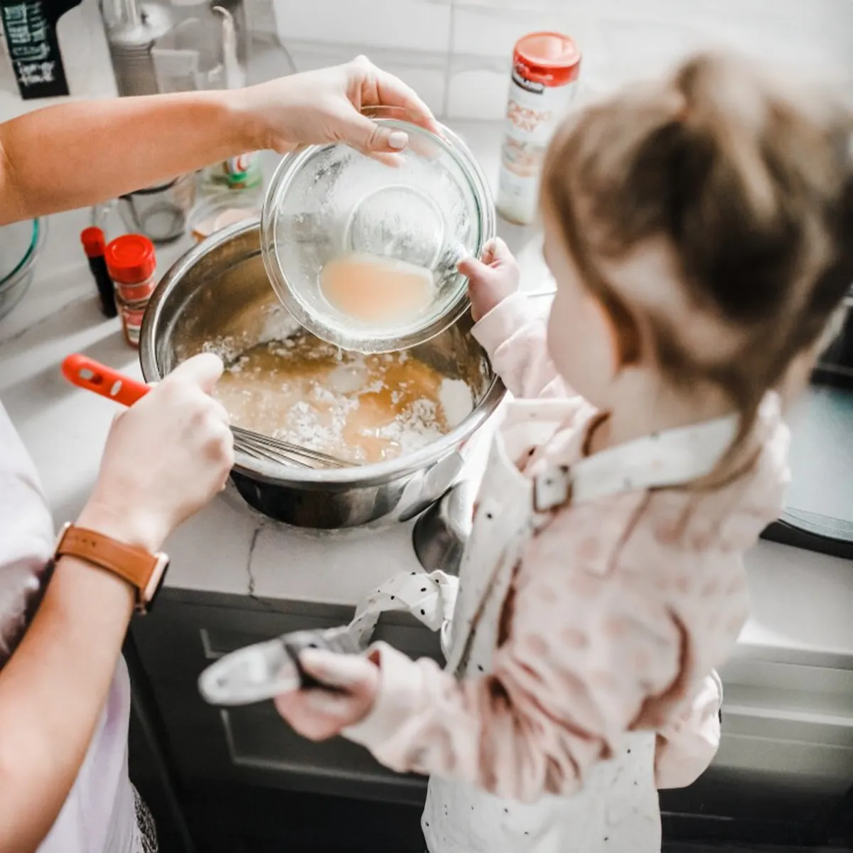 Adult and young child pour liquid into a mixing bowl while whisking batter together on a kitchen countertop