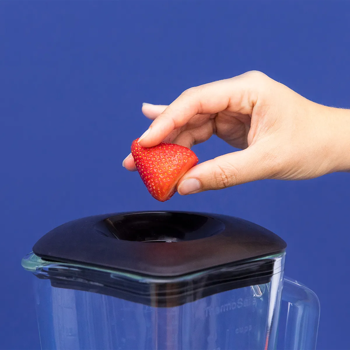 Hand holding a strawberry above a blender with a blue background.