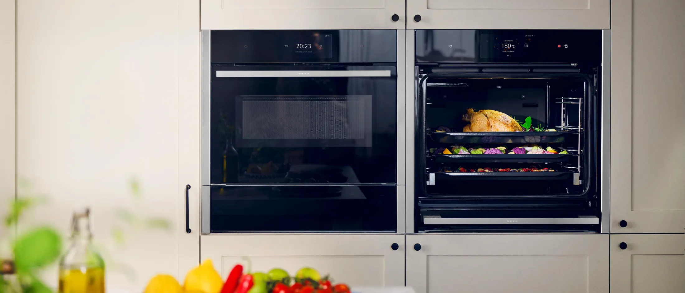 Dans une cuisine moderne, deux fours encastrés, l'un ouvert laissant apparaître un poulet rôti et des légumes, et des produits frais sur le plan de travail.