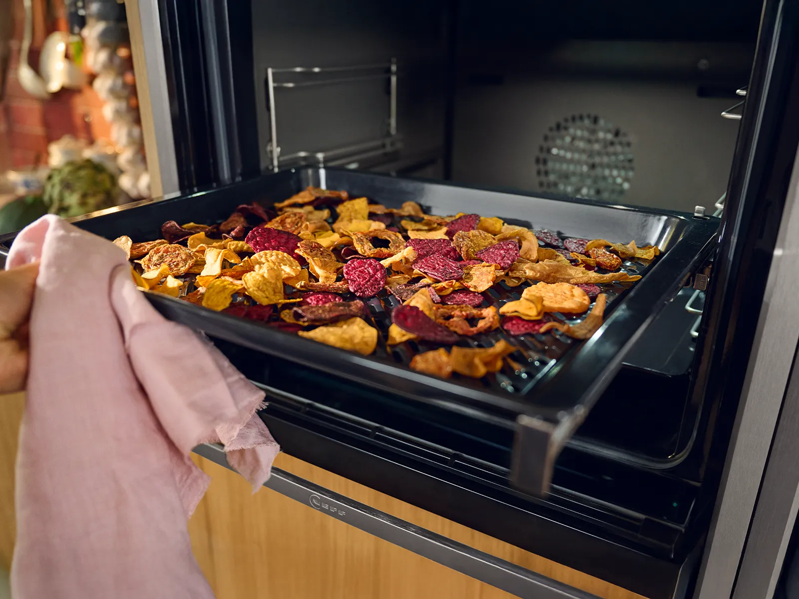 A hand wearing a pink cloth is pulling a tray of colorful, roasted vegetable chips out of an oven.