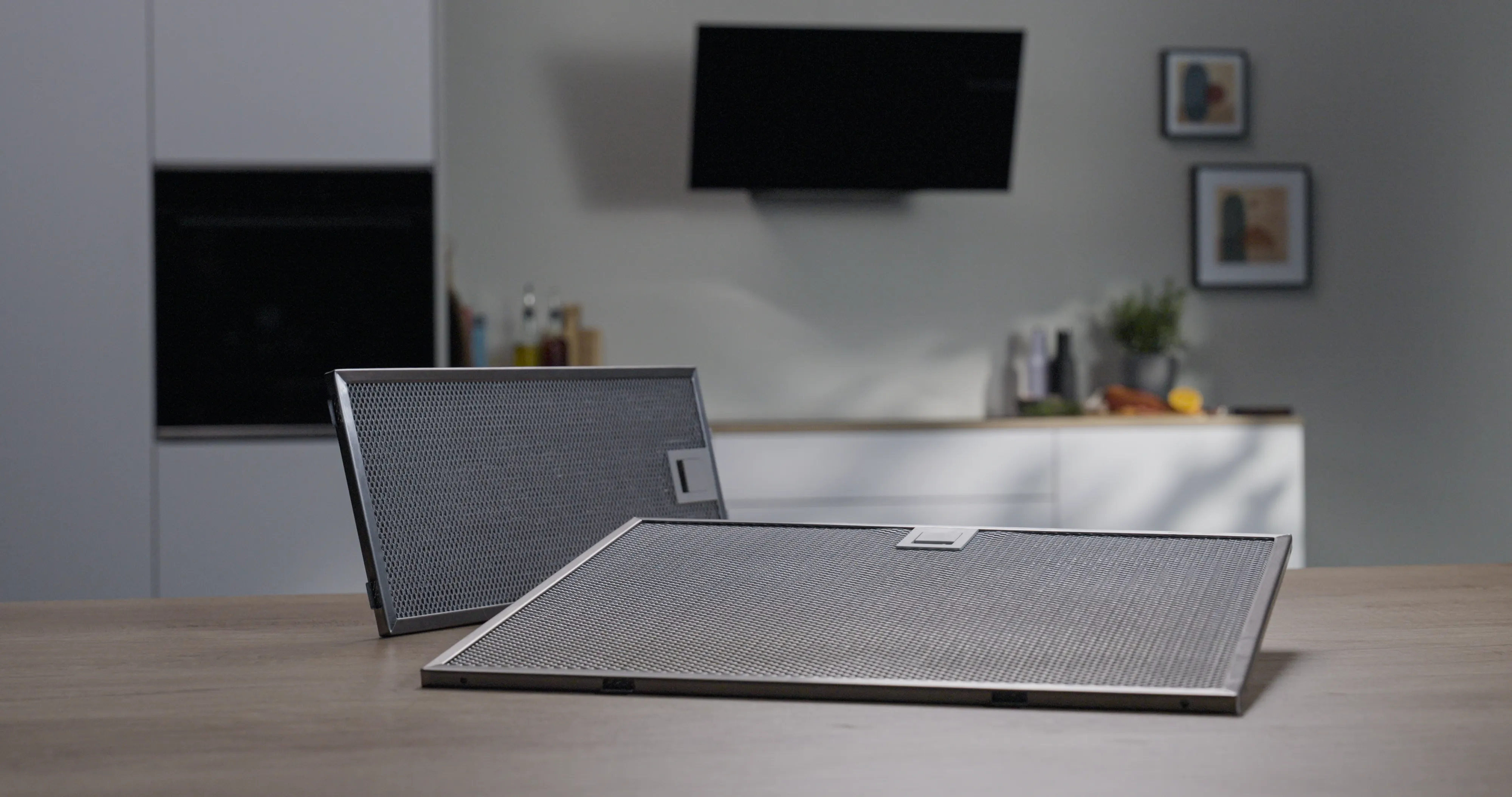 Zwei rechteckige Metallgitter-Fettfilter liegen auf einem Holztisch in einer modernen Küchenumgebung.