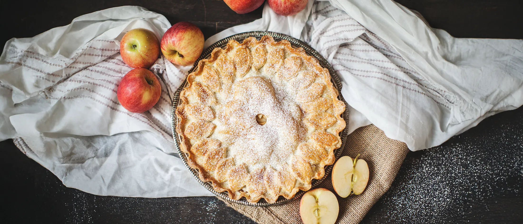 Baked apple pie with decorative crust and powdered sugar on a plate surrounded by whole and halved apples on cloths