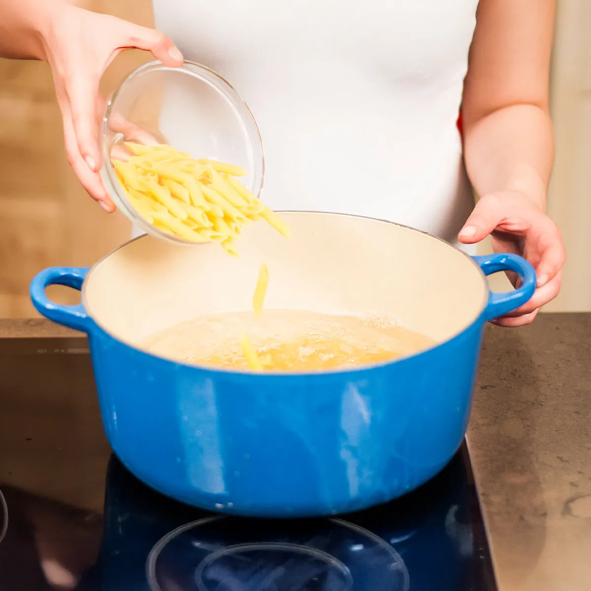 Person pouring penne pasta from a glass bowl into a large blue pot on a stovetop.