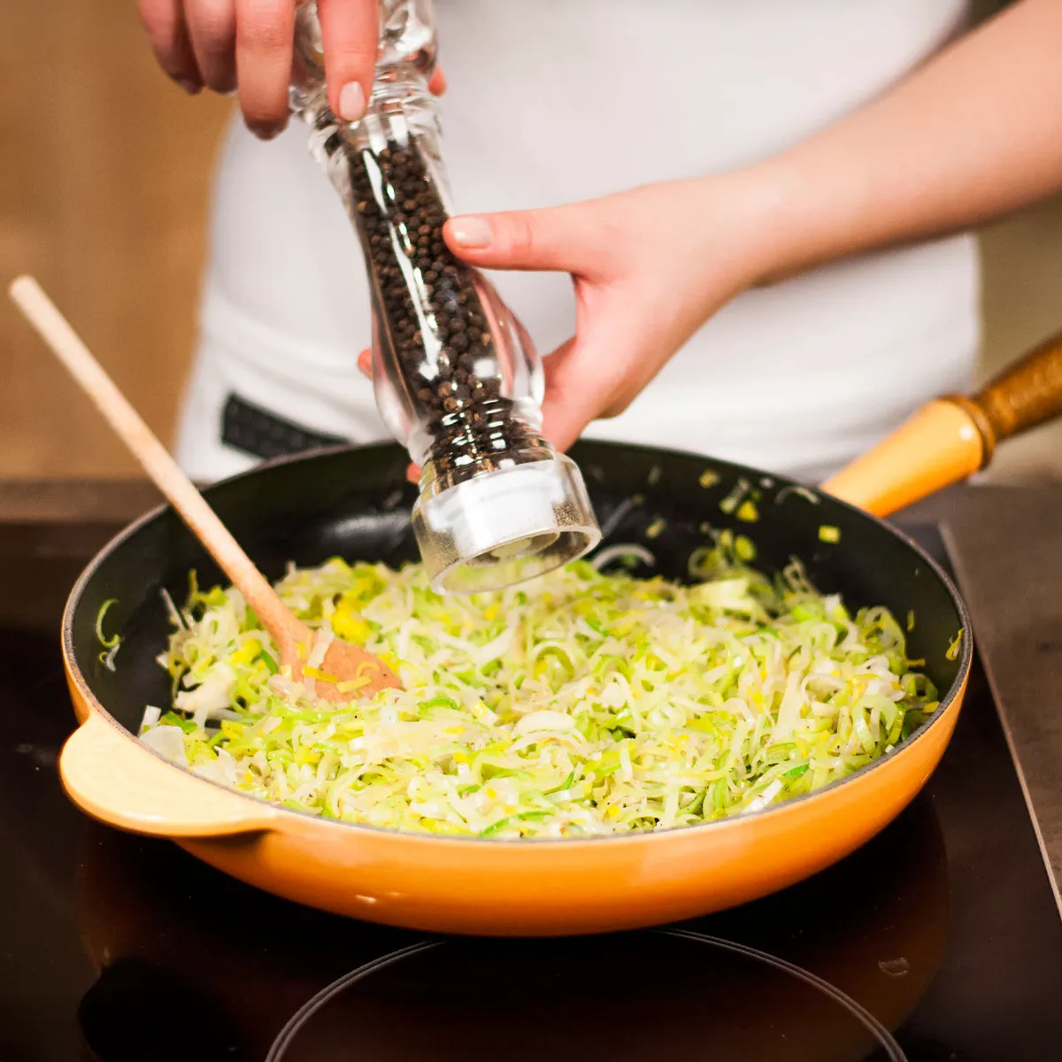 Hands seasoning chopped leeks in a frying pan with a pepper grinder.