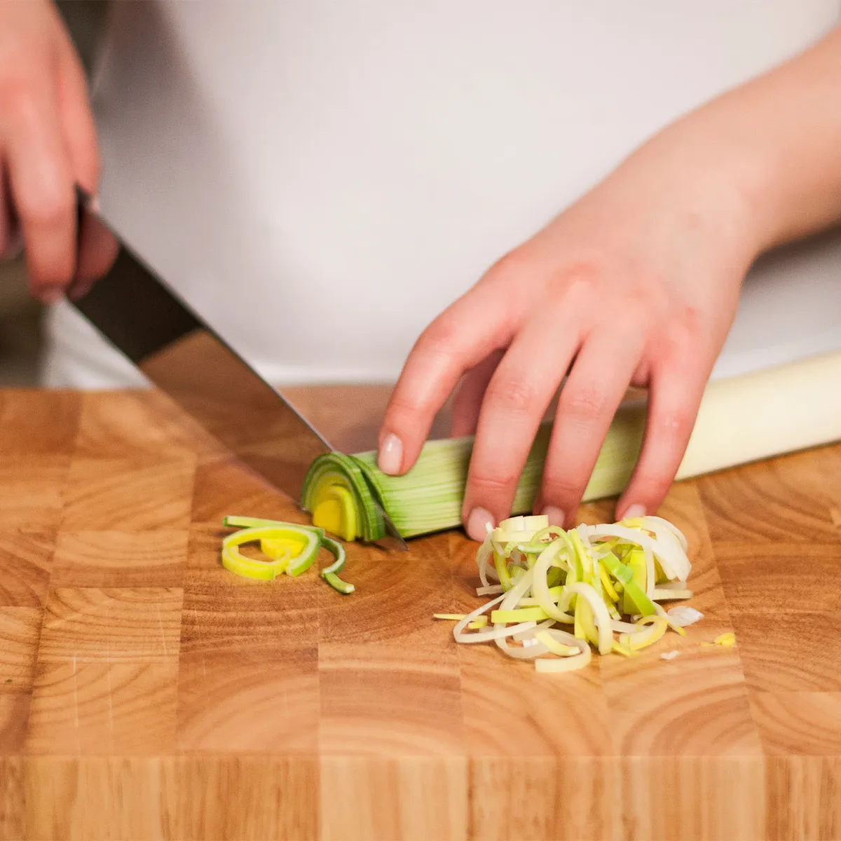 Hands slicing a leek into thin rings on a wooden cutting board.