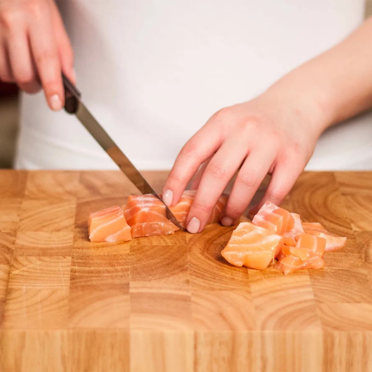 Hands cutting raw salmon into cubes on a wooden cutting board.