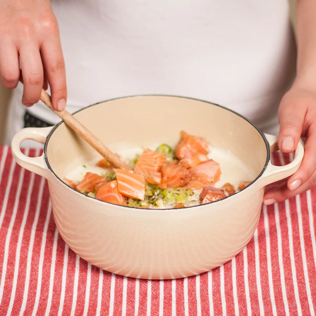 Hands stirring chunks of raw salmon and chopped leeks in a white ceramic pot on a red-and-white striped cloth.