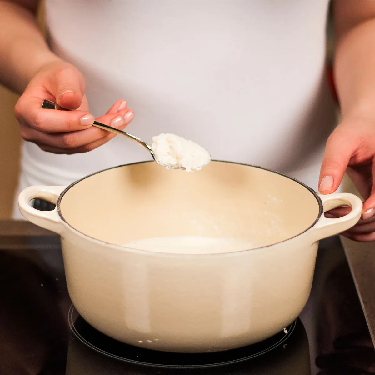 Person holding a spoon with a white powder above a cream-colored pot on a stovetop.