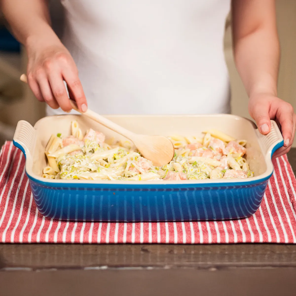 Hands stirring penne pasta with salmon and leeks in a blue ceramic baking dish on a red and white striped cloth.