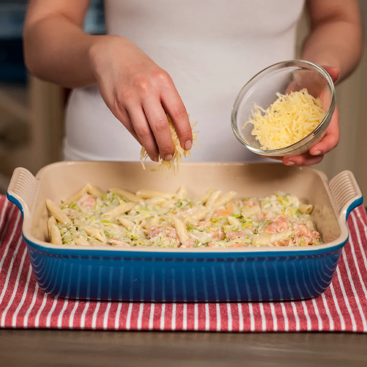 Hands sprinkling shredded cheese over a pasta casserole in a blue baking dish on a red and white striped cloth.