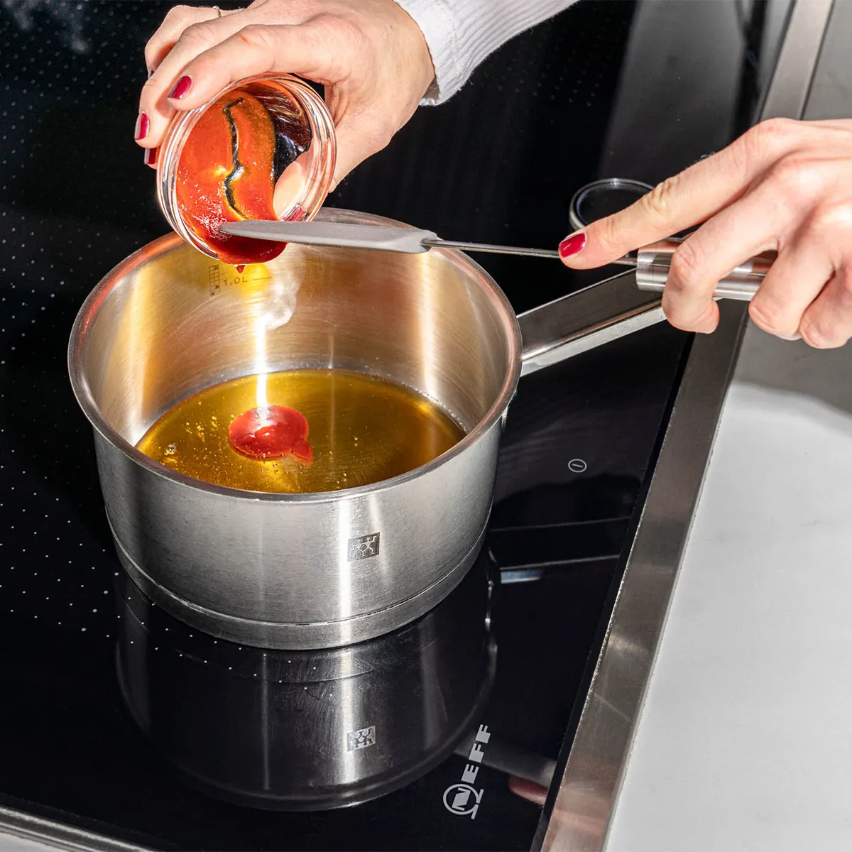Hands pouring red sauce from a jar into a small stainless-steel saucepan of oil on an induction cooktop.
