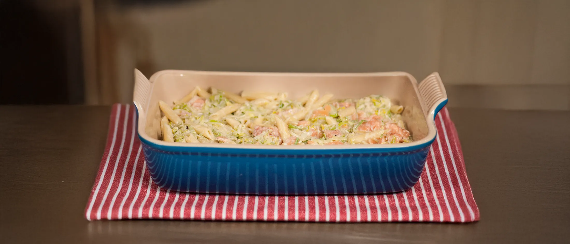 Blue rectangular baking dish with creamy pasta and vegetables on a red and white striped cloth on a wooden table.