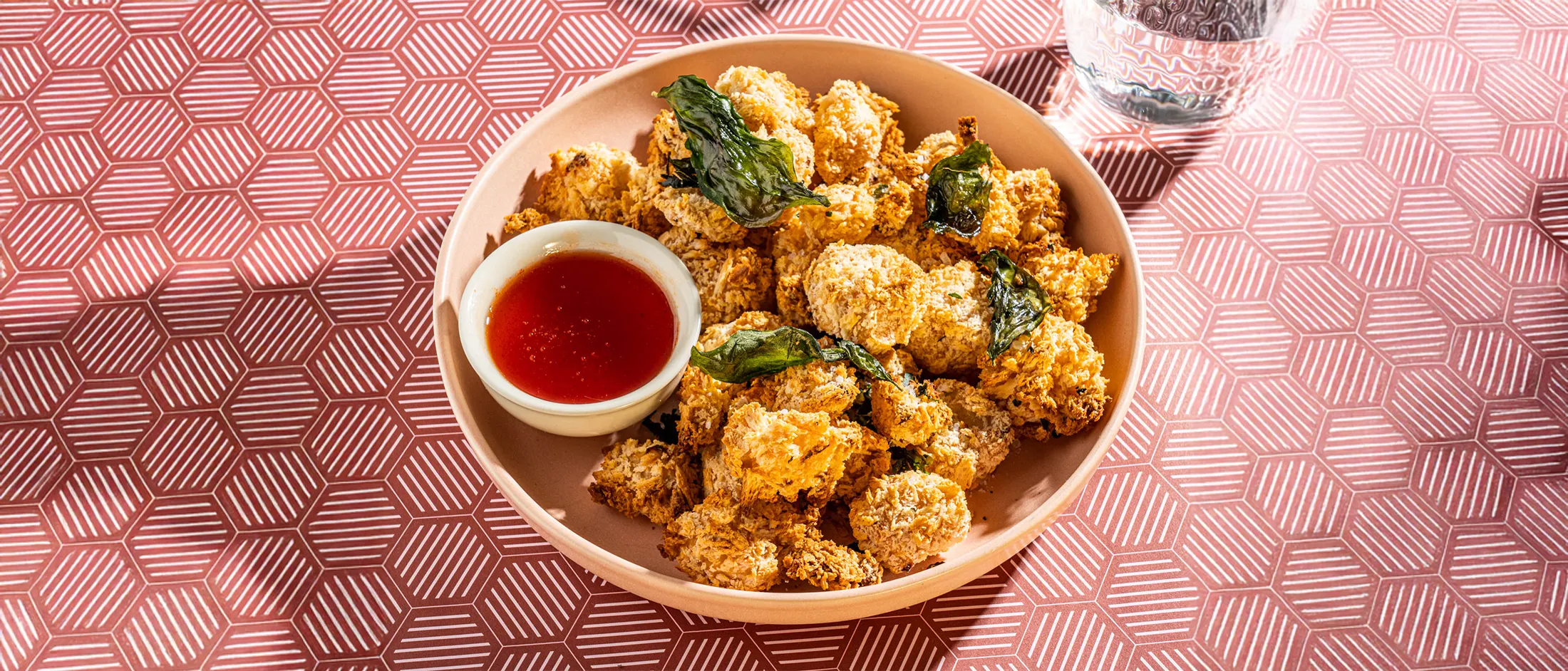 Close-up of a pink bowl filled with crispy golden breaded bites, garnished with fried basil leaves and a small dish of red dipping sauce on a patterned placemat.