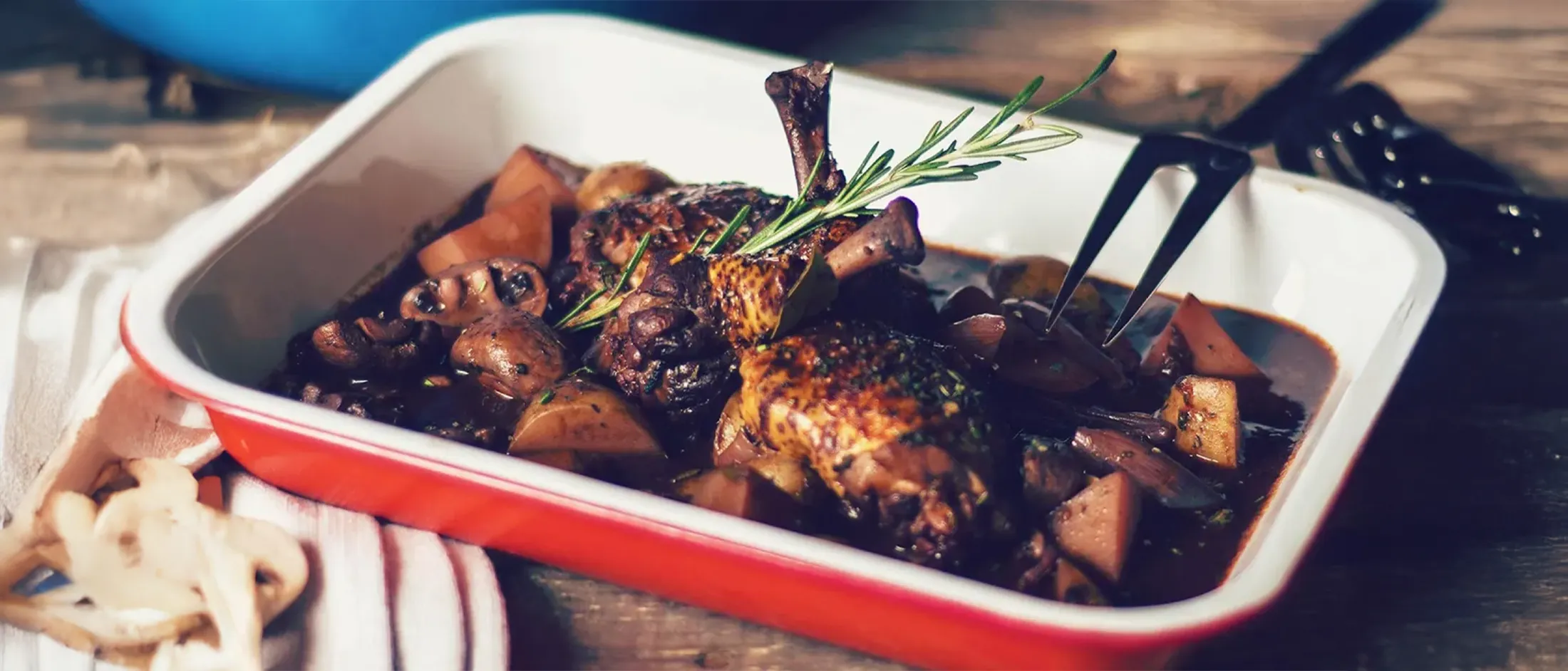 Braised chicken drumsticks with mushrooms, potatoes, and rosemary in a rich dark sauce in a red and white baking dish.
