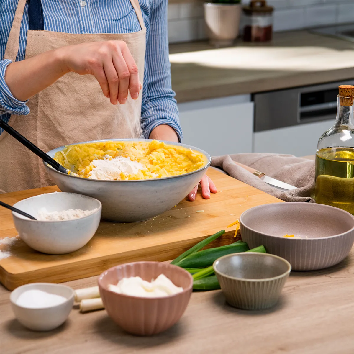 Person sprinkling flour into a bowl of mashed potatoes on a kitchen counter with various bowls and ingredients nearby.