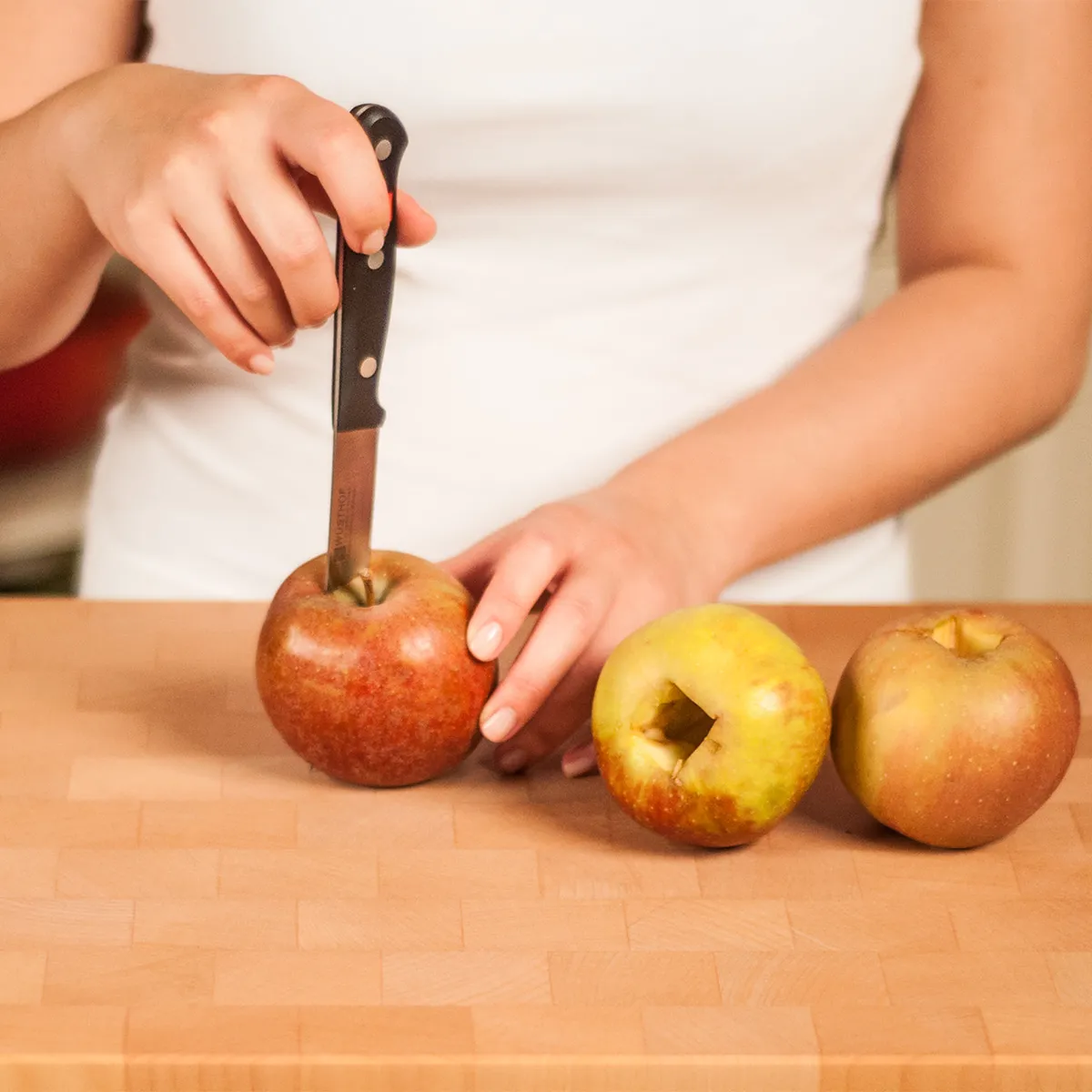 Person using a knife to core apples on a wooden cutting board with two cored apples already placed beside.