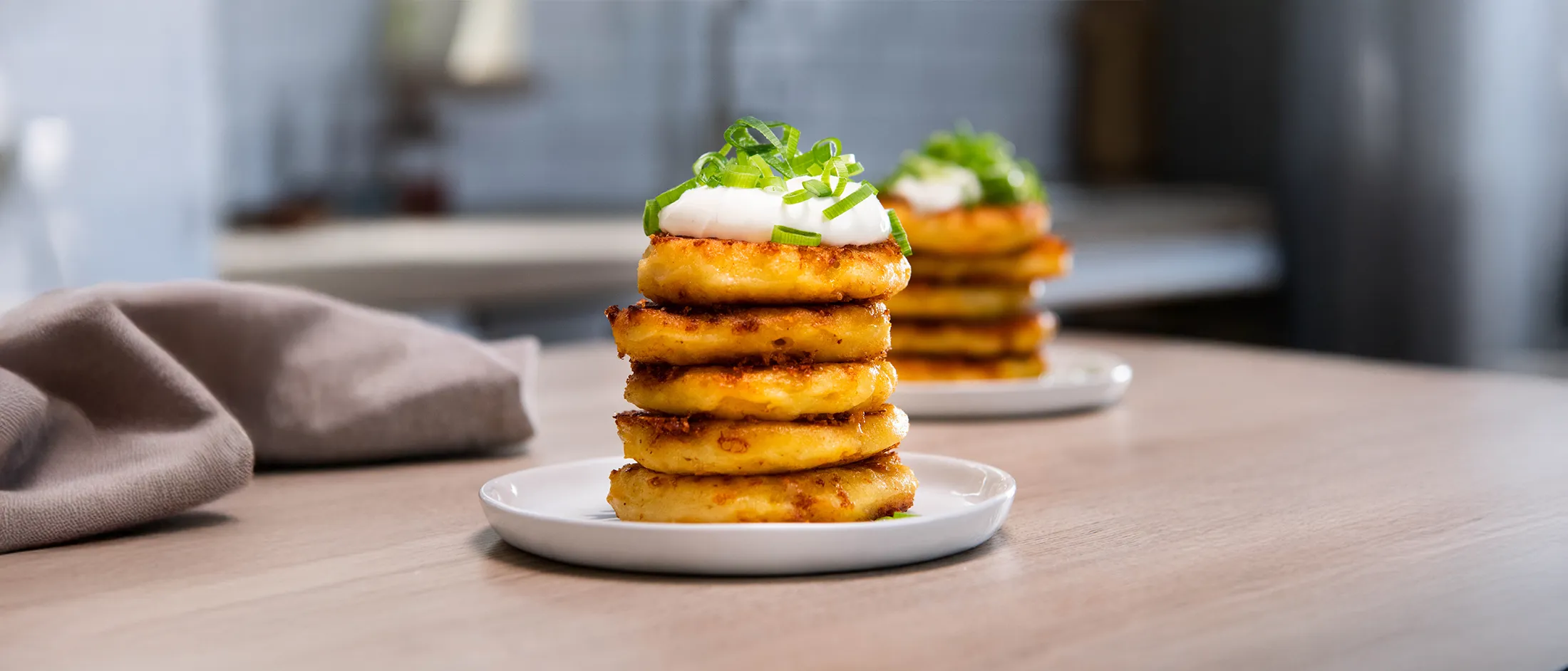 Stack of golden potato pancakes topped with sour cream and chopped green onions on a white plate, with a blurred background.