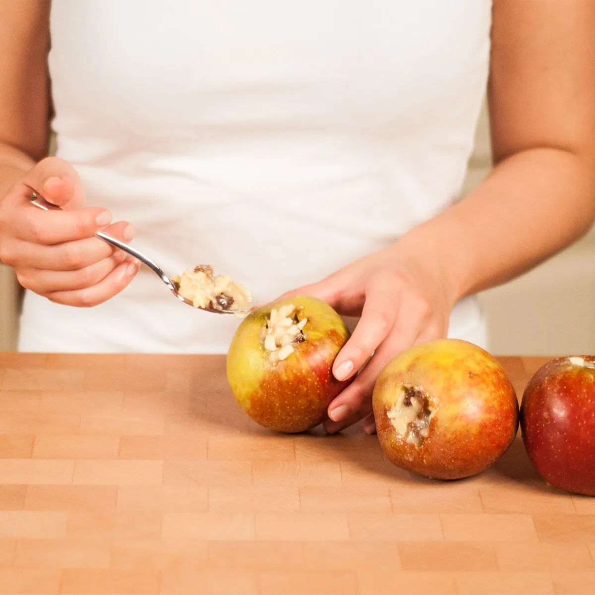 Person filling cored apples with a spoonful of oatmeal on a wooden countertop.