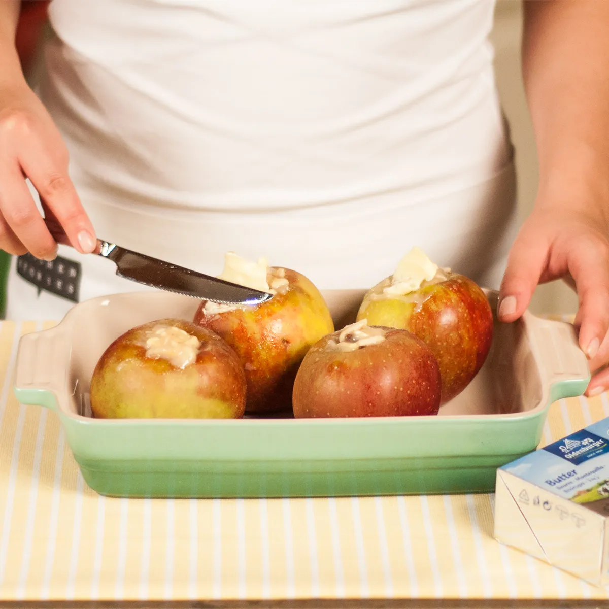 Close-up of a person in a white apron spreading pats of butter onto four marzipan-stuffed apples arranged in a green ceramic baking dish on a yellow-striped