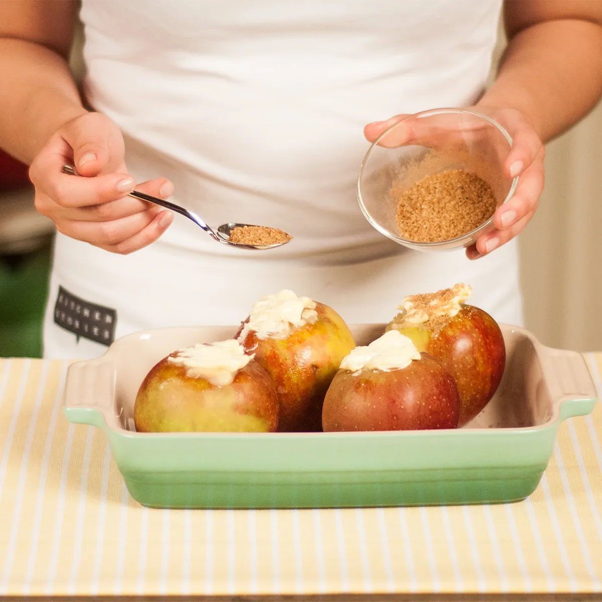 Close-up of a person in a white apron sprinkling brown sugar from a small glass bowl onto marzipan‑filled apples in a green ceramic baking dish set on a yellow‑striped tablecloth.