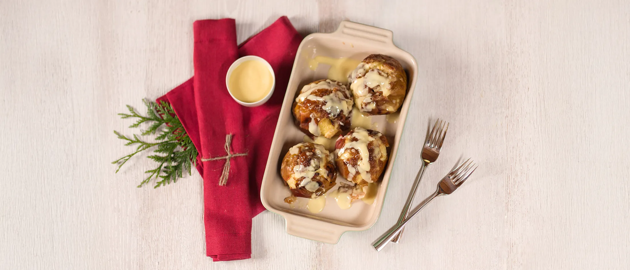 Overhead shot of a beige rectangular baking dish holding four warm, sticky glazed buns drizzled with icing on a pale wooden table; a folded red napkin tied with twine and a small ramekin with an evergreen sprig sit to the left and two forks rest crossed to the right.