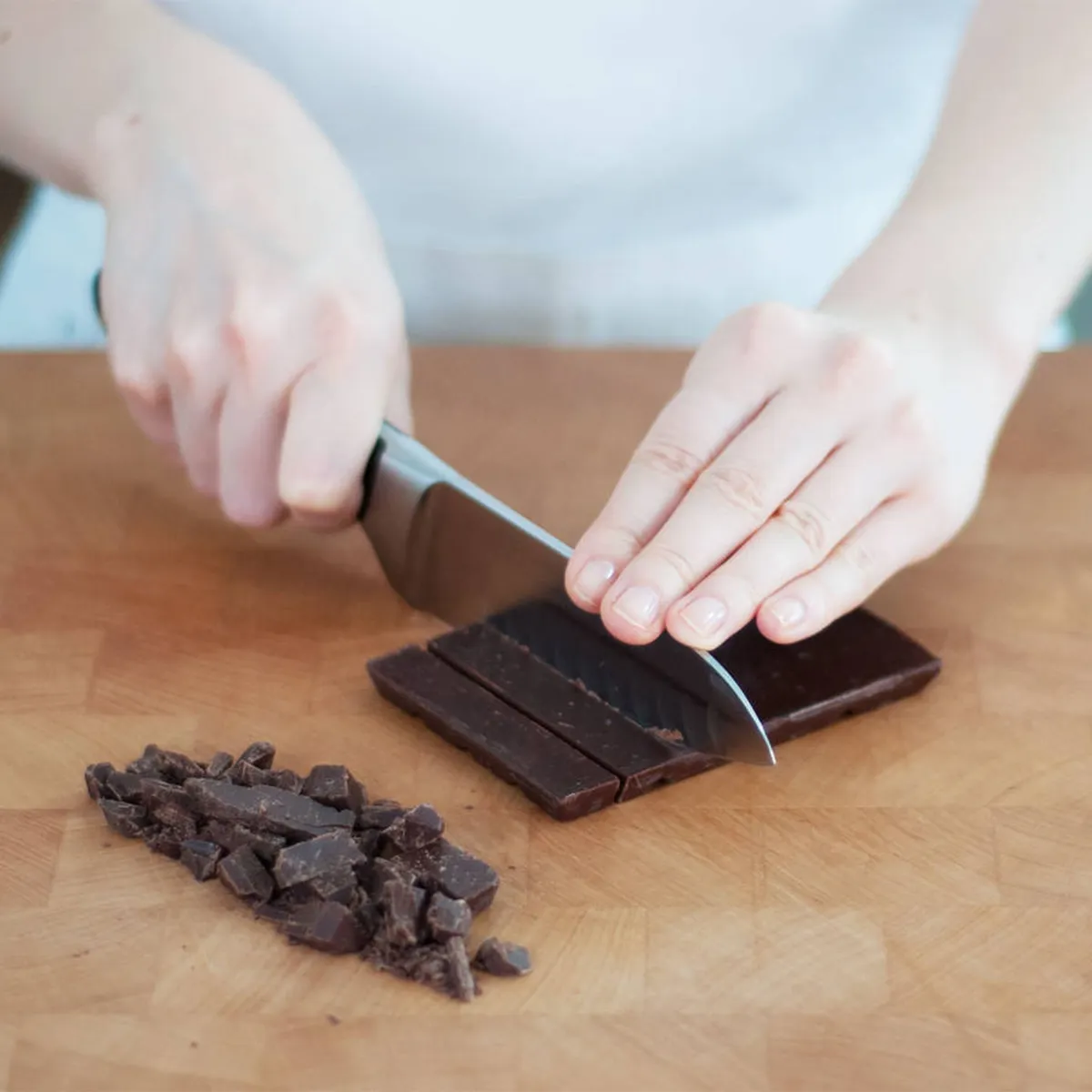 Person slicing dark chocolate on a wooden cutting board with a knife, with chopped pieces beside it