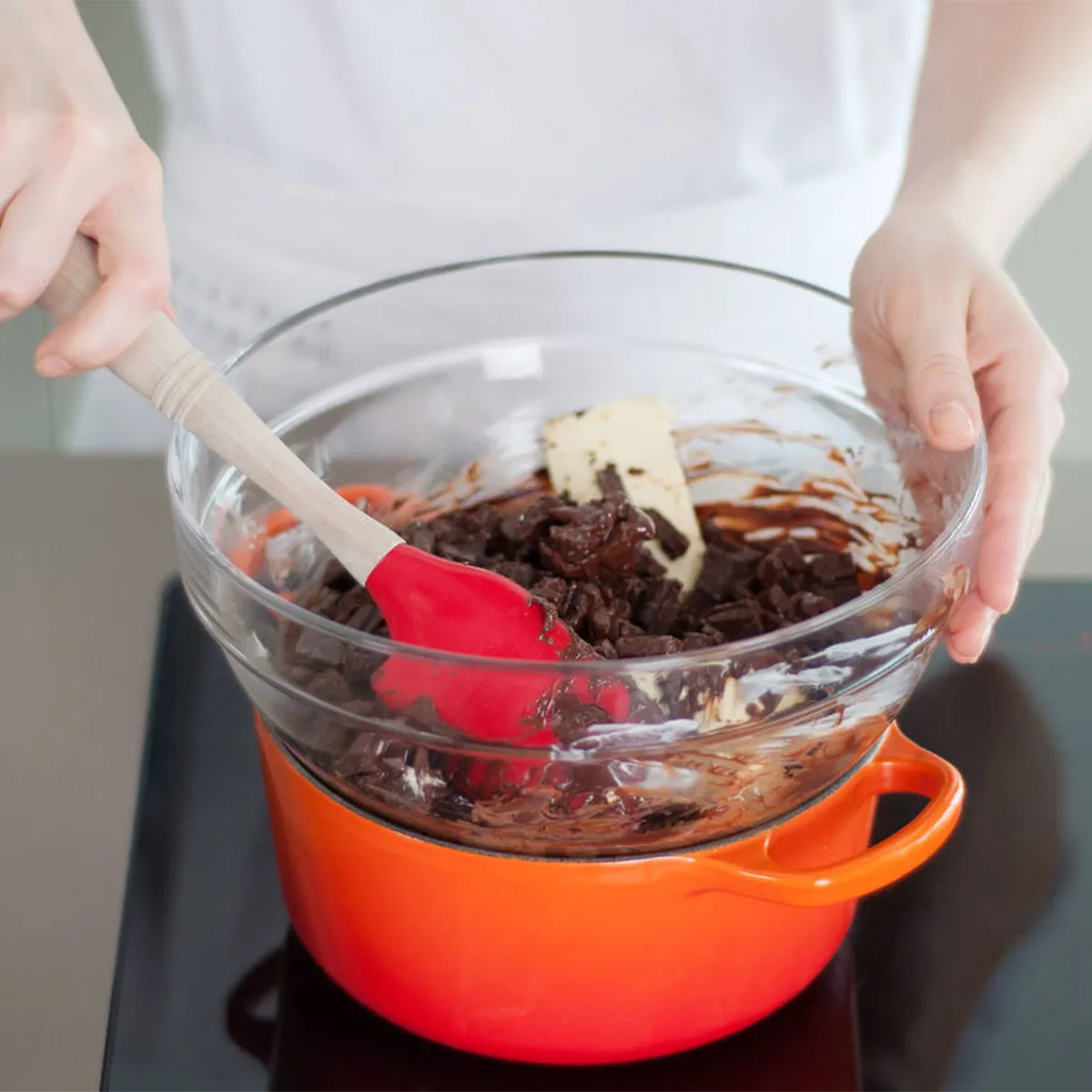 Person stirring chocolate chunks in a glass bowl set over an orange pot on a stovetop, melting chocolate using a double boiler method