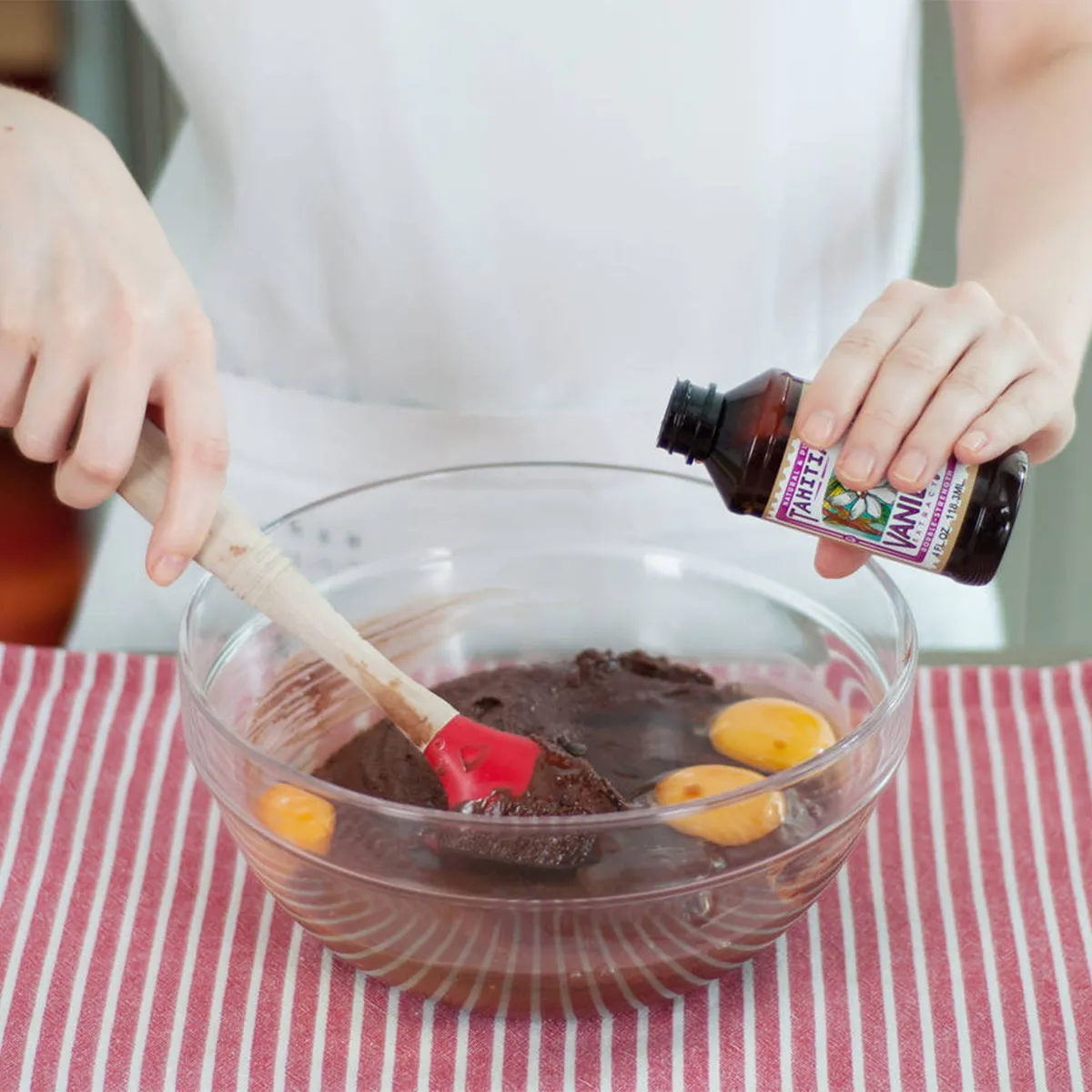 Person adding vanilla extract to a glass bowl with eggs and chocolate batter, stirring with a red spatula on a striped cloth