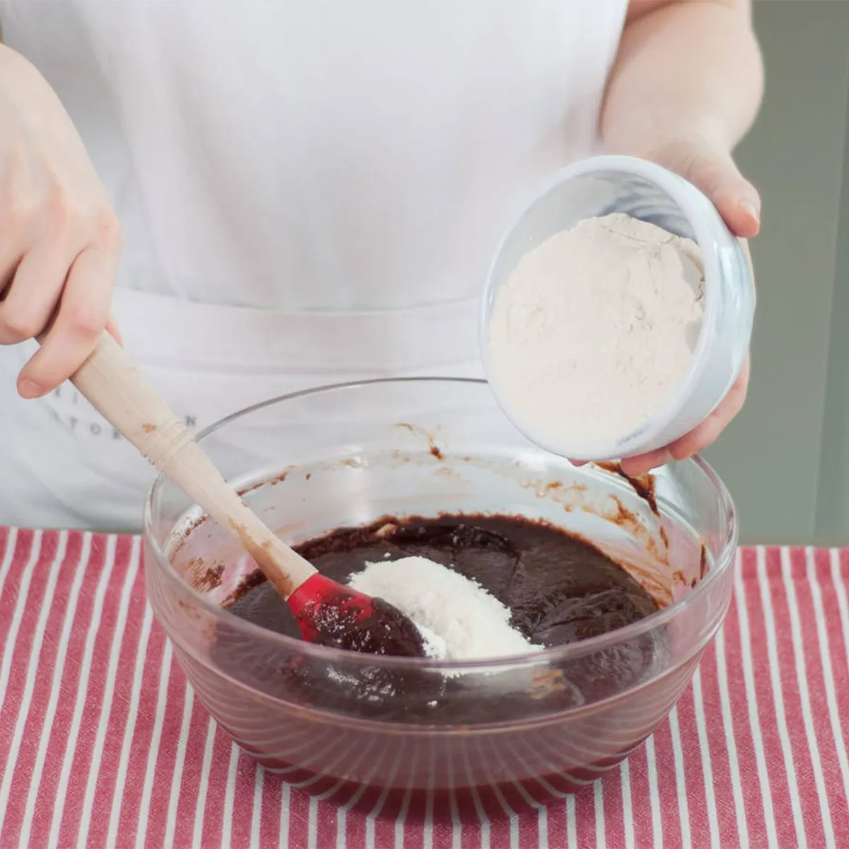 Person folding flour into chocolate batter before transferring it to a lined baking pan for brownies