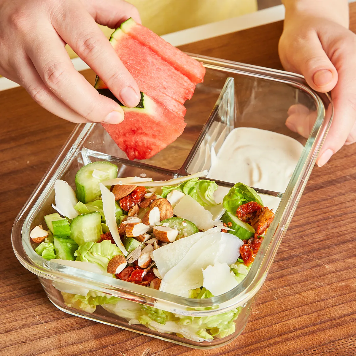 Close-up of hands placing a slice of watermelon into a divided glass meal-prep container on a wooden countertop; one compartment holds a green salad with