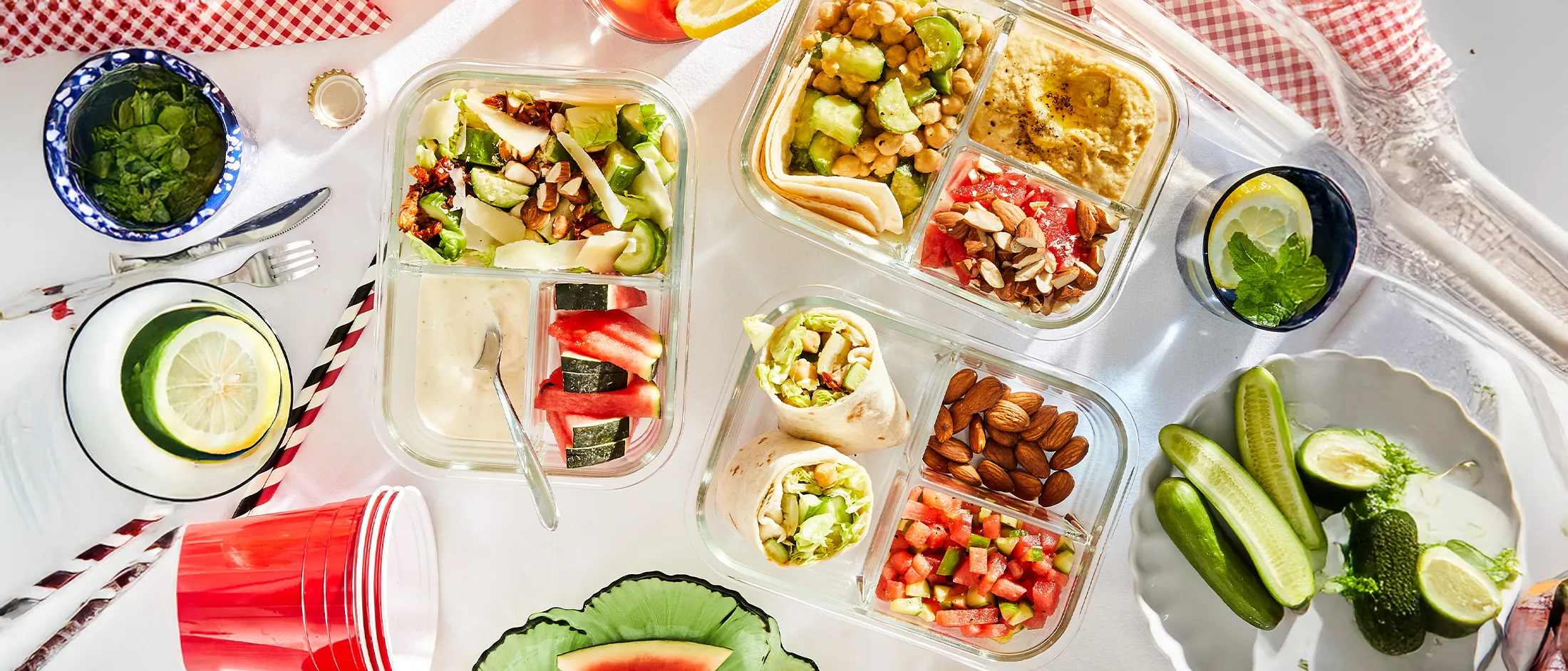 Picnic spread with salads, wraps, hummus, watermelon slices, almonds, cucumbers, and lemon drinks on a white tablecloth with red checkered napkins.