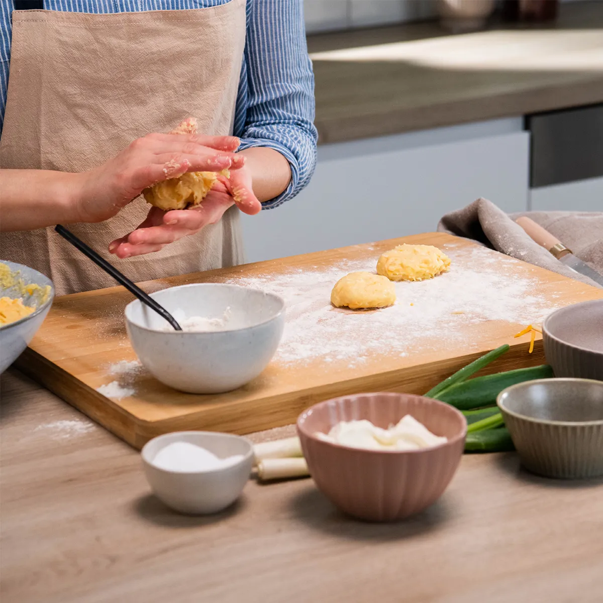 Person shaping dough balls on a floured wooden board with bowls of ingredients on a kitchen counter.