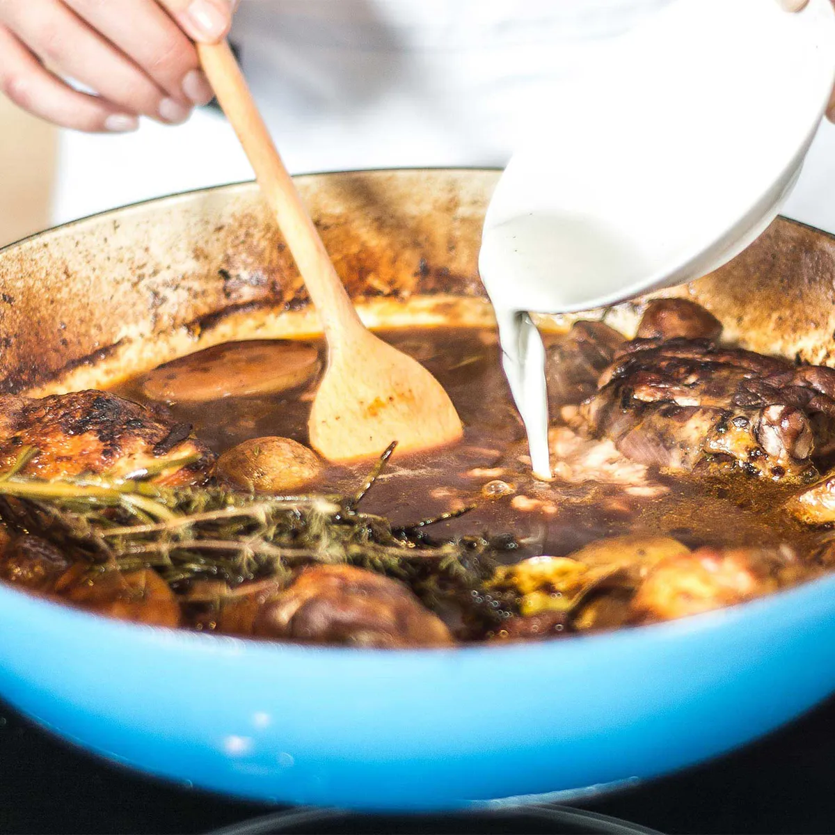 Close-up of hands pouring cream into a blue enameled casserole with browned chicken thighs, potatoes, mushrooms and sprigs of thyme; a wooden spoon is in the stew.