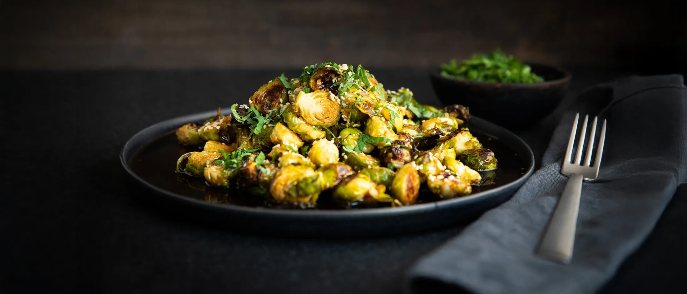 Plate of roasted Brussels sprouts garnished with chopped herbs on a dark surface with a fork and napkin beside it