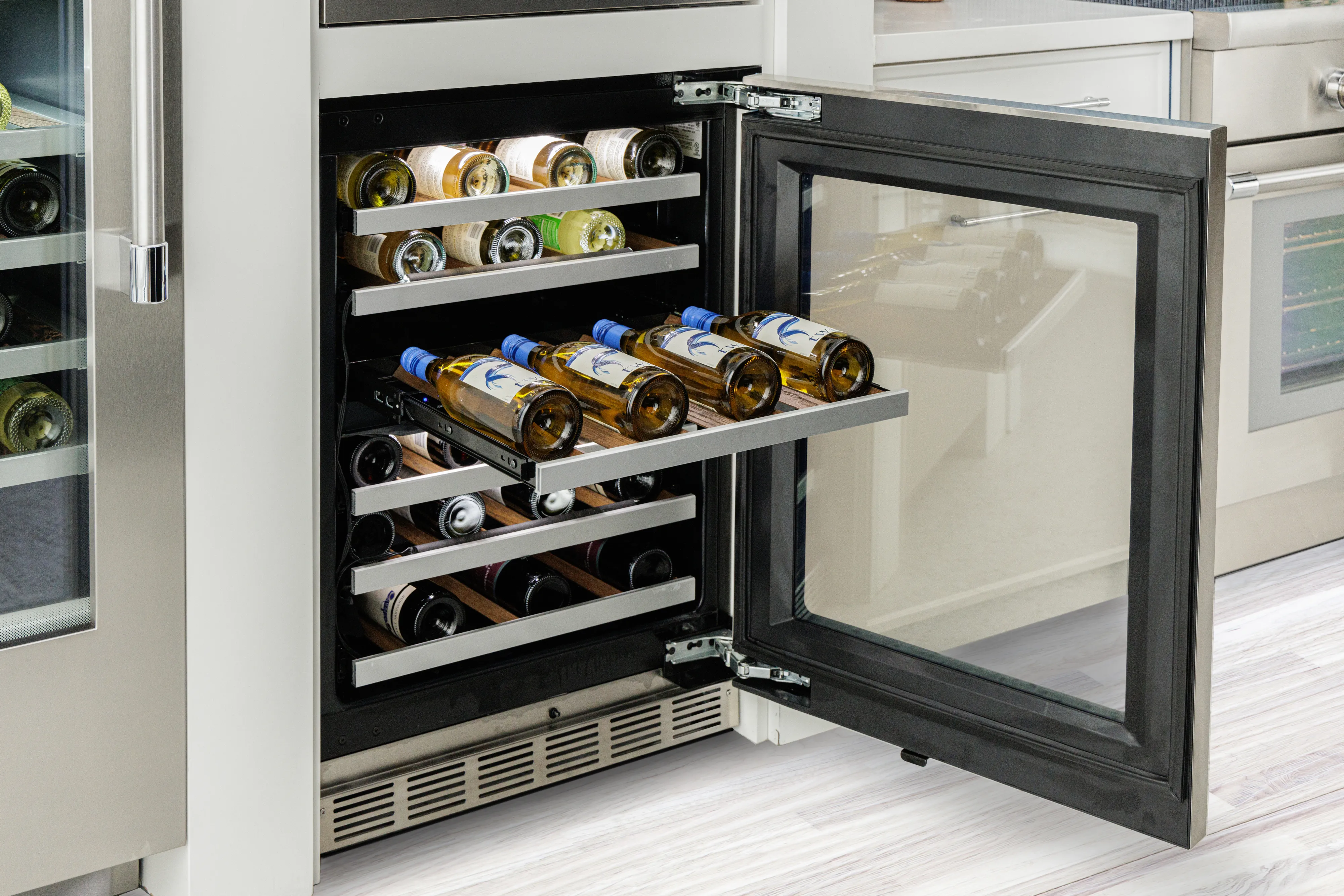 Open under-counter wine cooler with multiple wine bottles stored on sliding shelves in a modern kitchen setting.