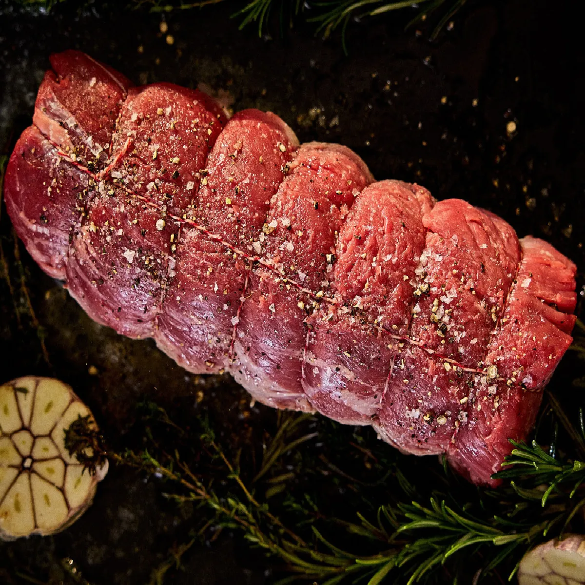 Affettato bistecca di manzo crudo con rosmarino, aglio e condimento su uno sfondo scuro.