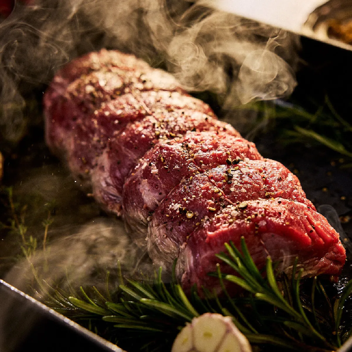 Bistecca con rosmarino, aglio e fumo su un vassoio di metallo scuro.