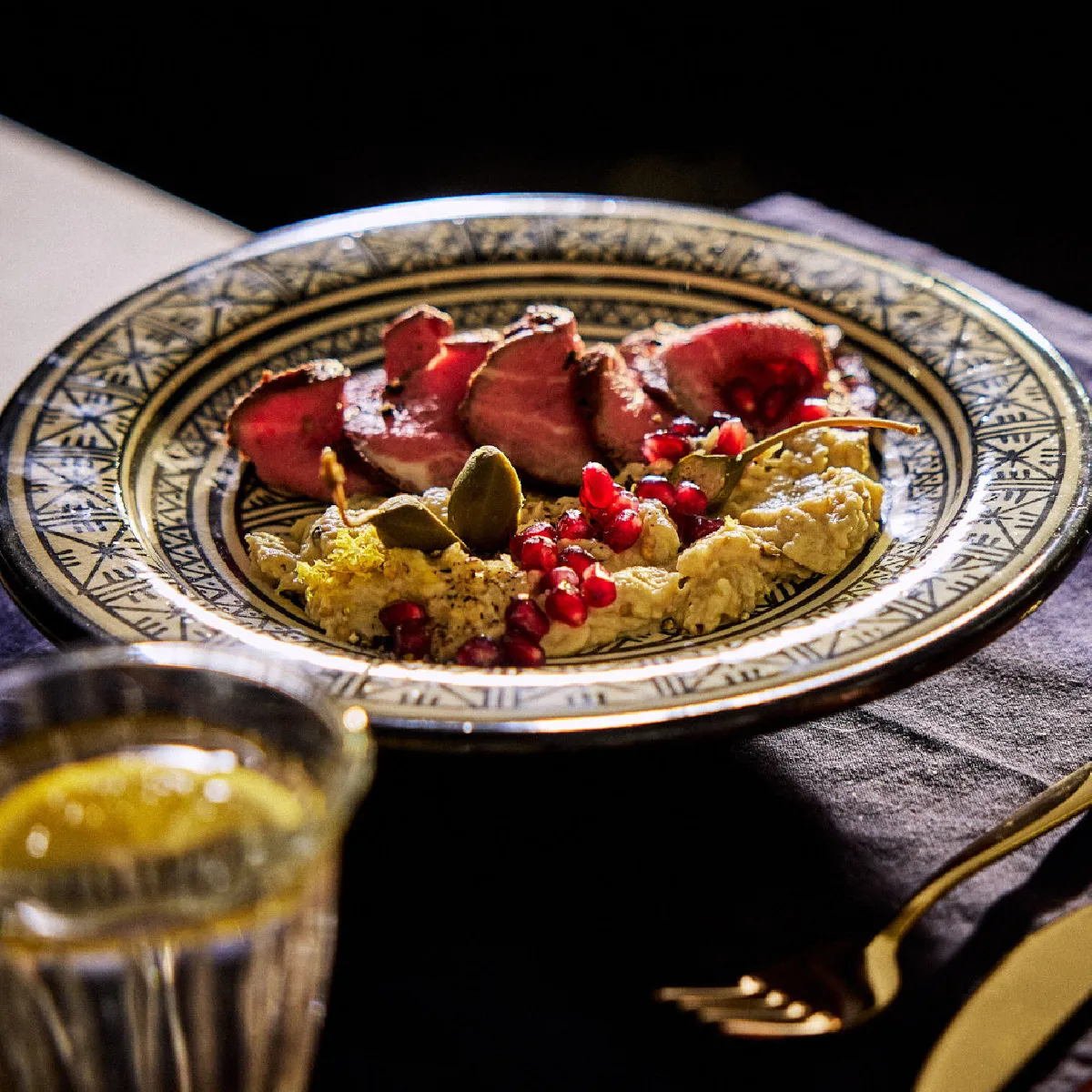 Piastra ornata con verdure arrosto, semi di melograno e utensili su uno sfondo di stoffa scuro.