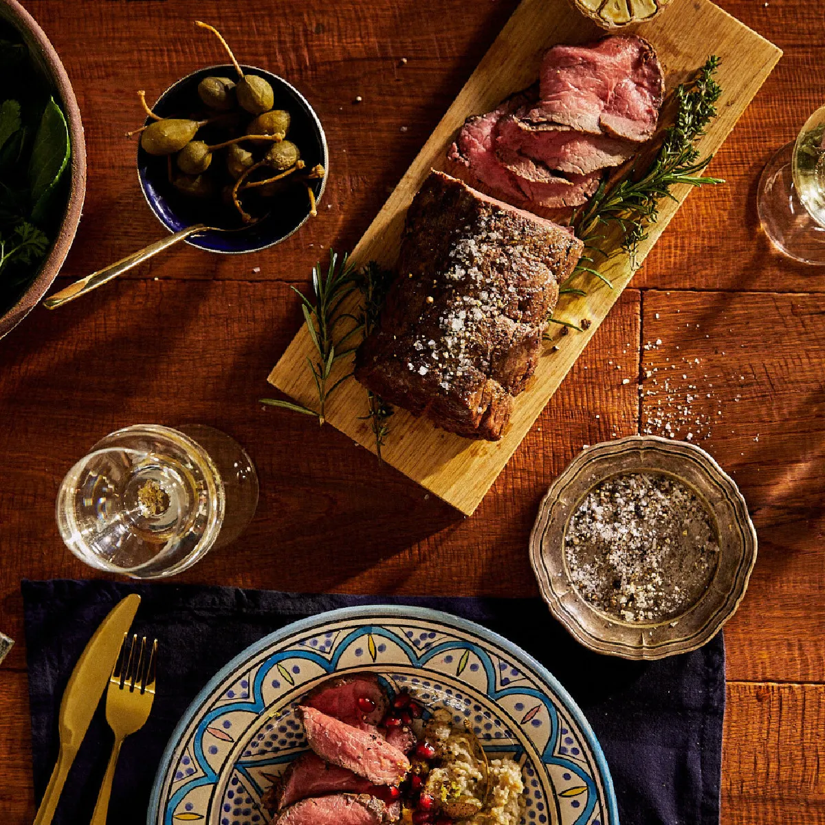Tavolo rustico con carne alla griglia, insalata, olive e bicchieri di vino su una superficie di legno.