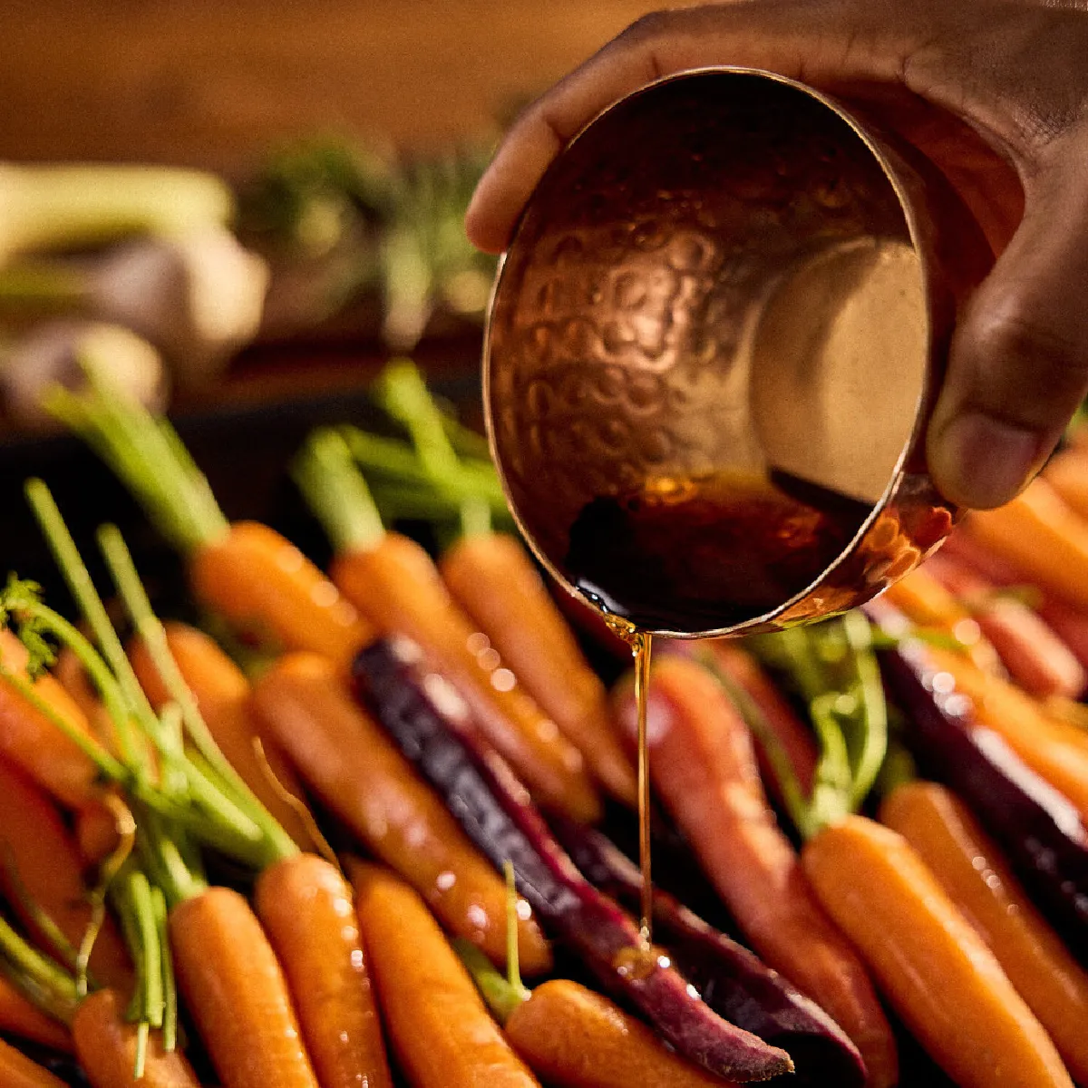 Mano che versa una miscela di acqua e sciroppo d'acero da un piccolo recipiente su una teglia di carote multicolori arrostite, con erbe aromatiche sullo sfondo.