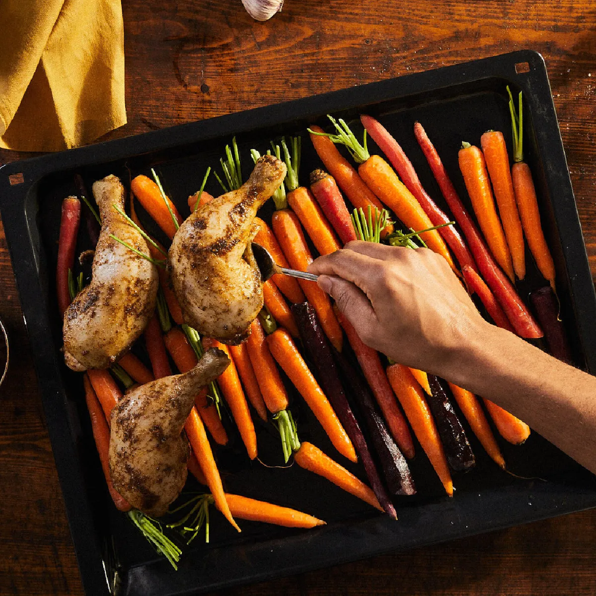 Mano che sistema erbe su una teglia con cosce di pollo arrosto e carote multicolori, accompagnata da patate novelle e un recipiente di salsa su un tavolo di legno.
