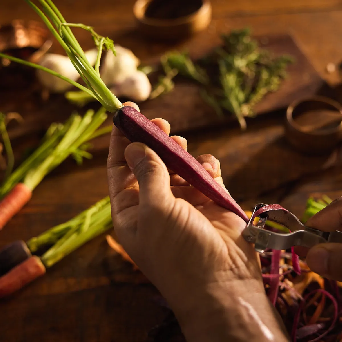 Mani che sbucciano una carota viola con un pelapatate su un tagliere di legno rustico, con altre carote colorate, aglio, erbe e patate novelle sfocate sullo sfondo.
