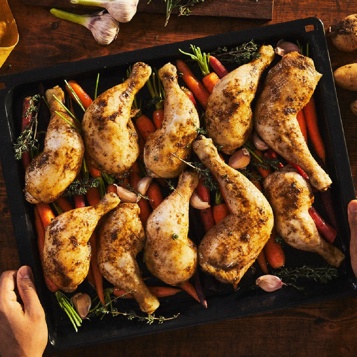Mani che sollevano una teglia con cosce di pollo arrosto, carote multicolori e erbe aromatiche, accompagnata da patate novelle e una ciotola di salsa su un tavolo rustico.