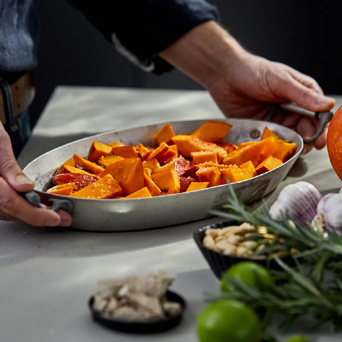 Una persona che tiene una teglia con pezzi di zucca affettata sopra il piano di lavoro della cucina, con erbe aromatiche, aglio e una zucca intera nelle vicinanze.